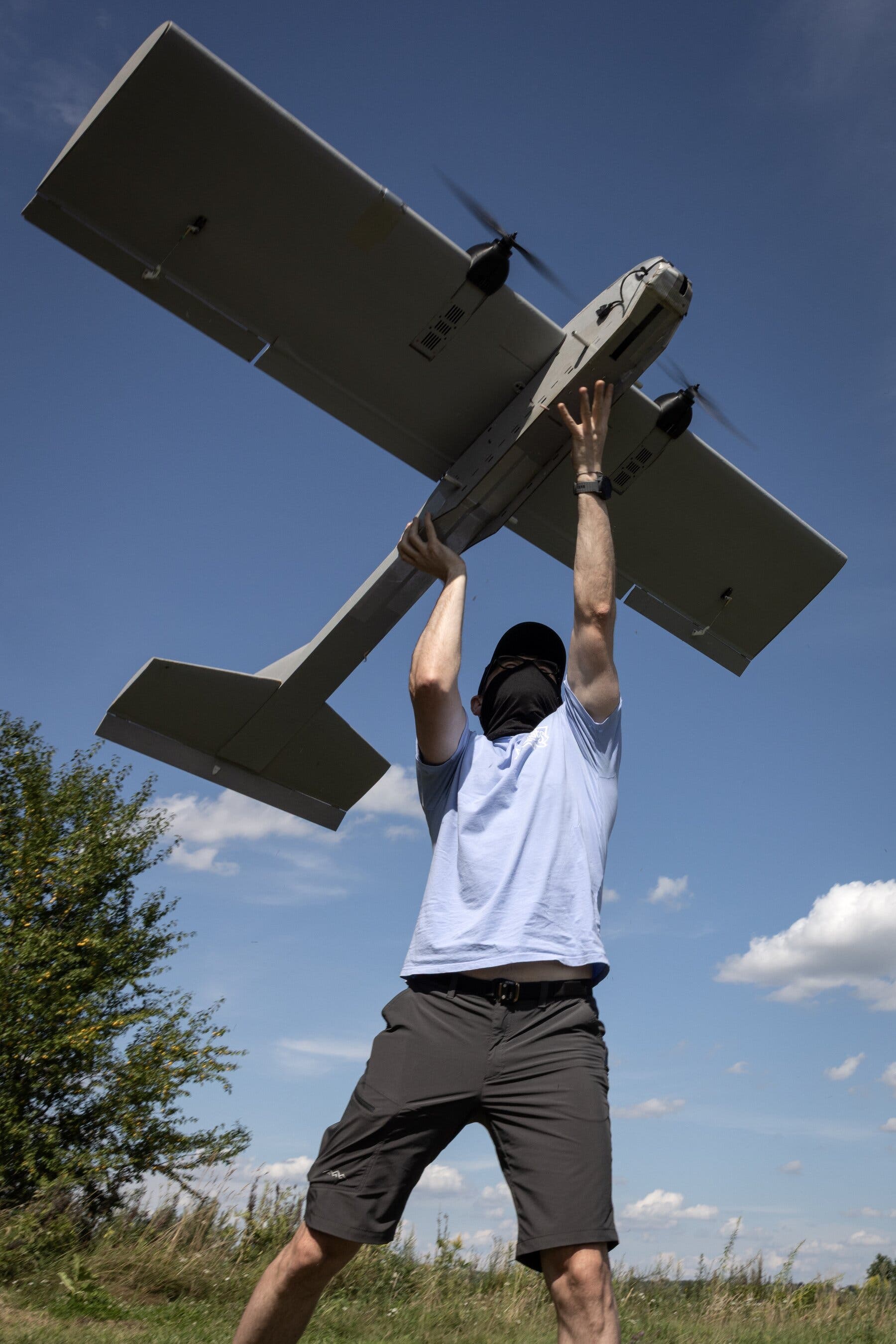 2026-01-05 ¡15mag-aidrone-1-mobileMasterAt3x.jpg|A masked man in a T-shirt and shorts stands in a green field under a blue sky holding an airplane-shaped drone above his head.