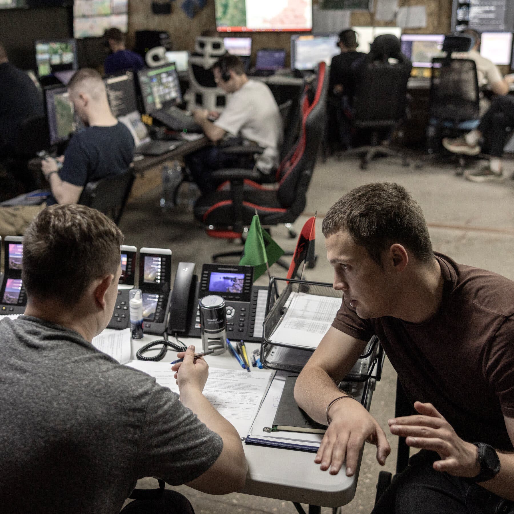 2026-01-05 ¡25mag-aidrones-09-mobileMasterAt3x.jpg|Two men talking closely in an improvised war command center with computers, maps, screens, clocks, and other people working at desks around them. 