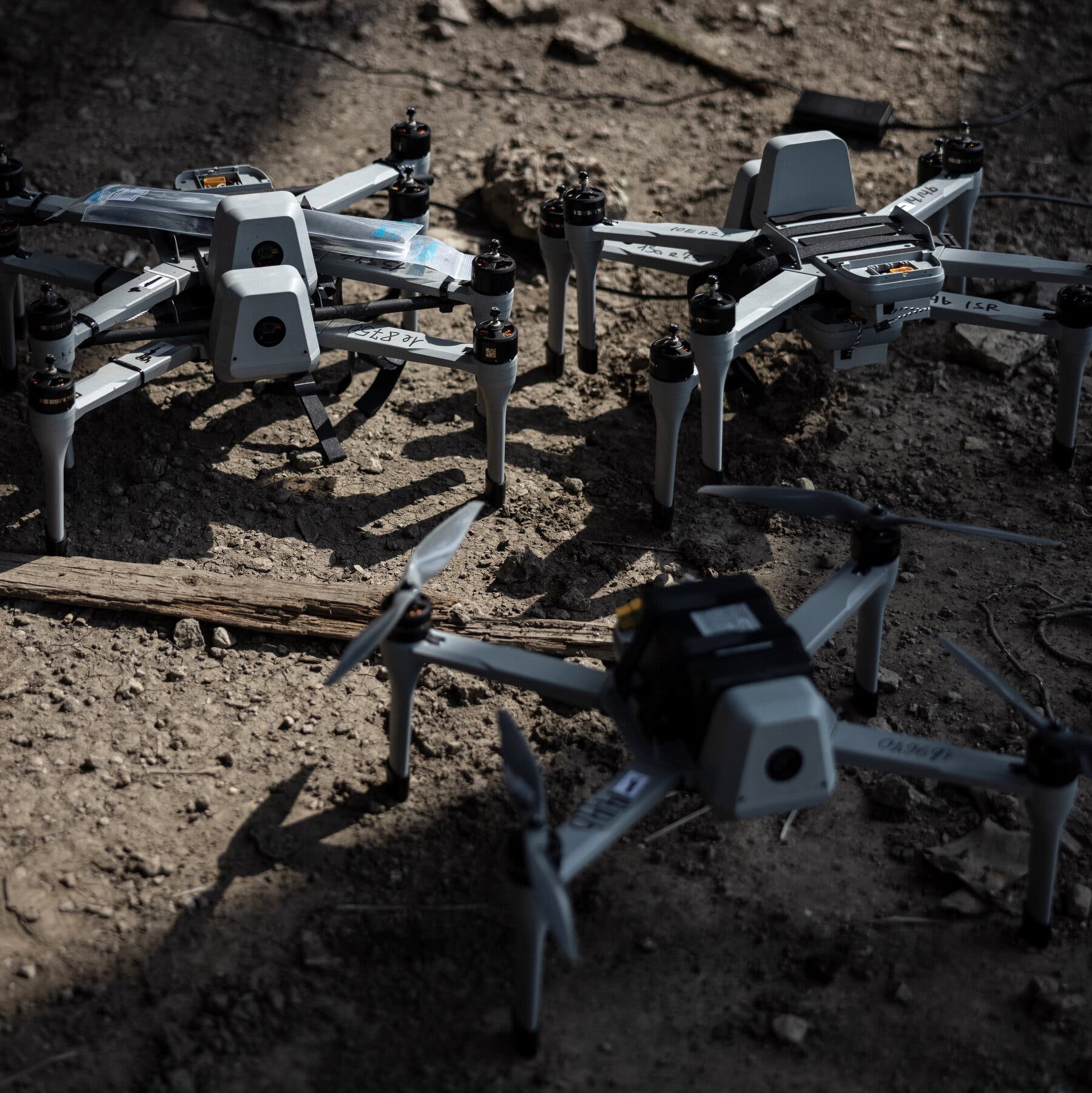 2026-01-05 ¡25mag-aidrones-mobileMasterAt3x.jpg|Three small military attack drones sit on a patch of dirt with a splash of sunlight highlighting the middle of the frame.