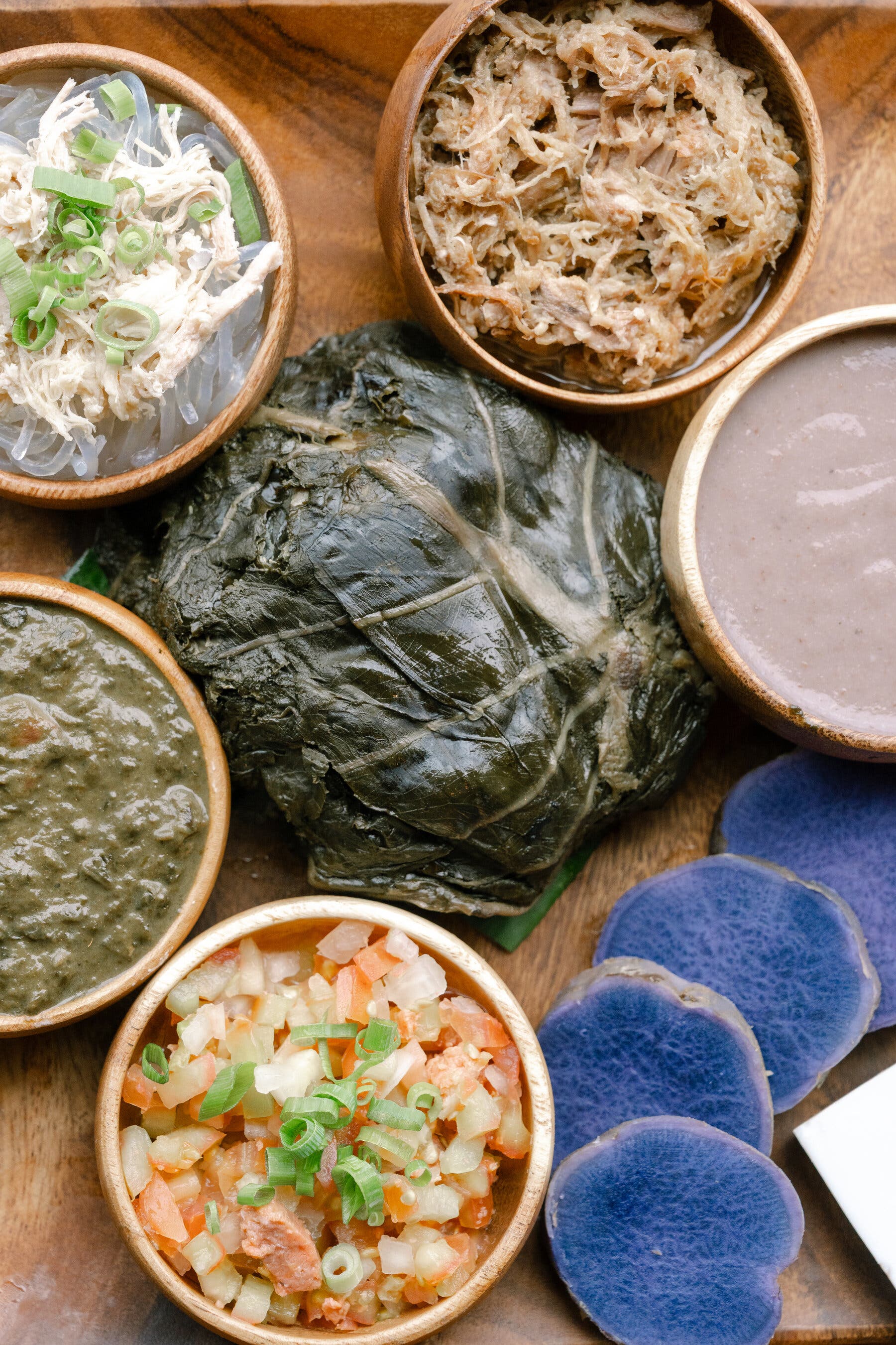 2026-02-28 ¡27trav-cheapskate-honolulu-bfkt-mobileMasterAt3x.jpgAn overhead view of a large, dark green leaf-wrapped dish and several small wooden bowls containing different colorful foods. These include shredded white meat with noodles, shredded brown meat, purple taro slices, and two kinds of pureed dishes.