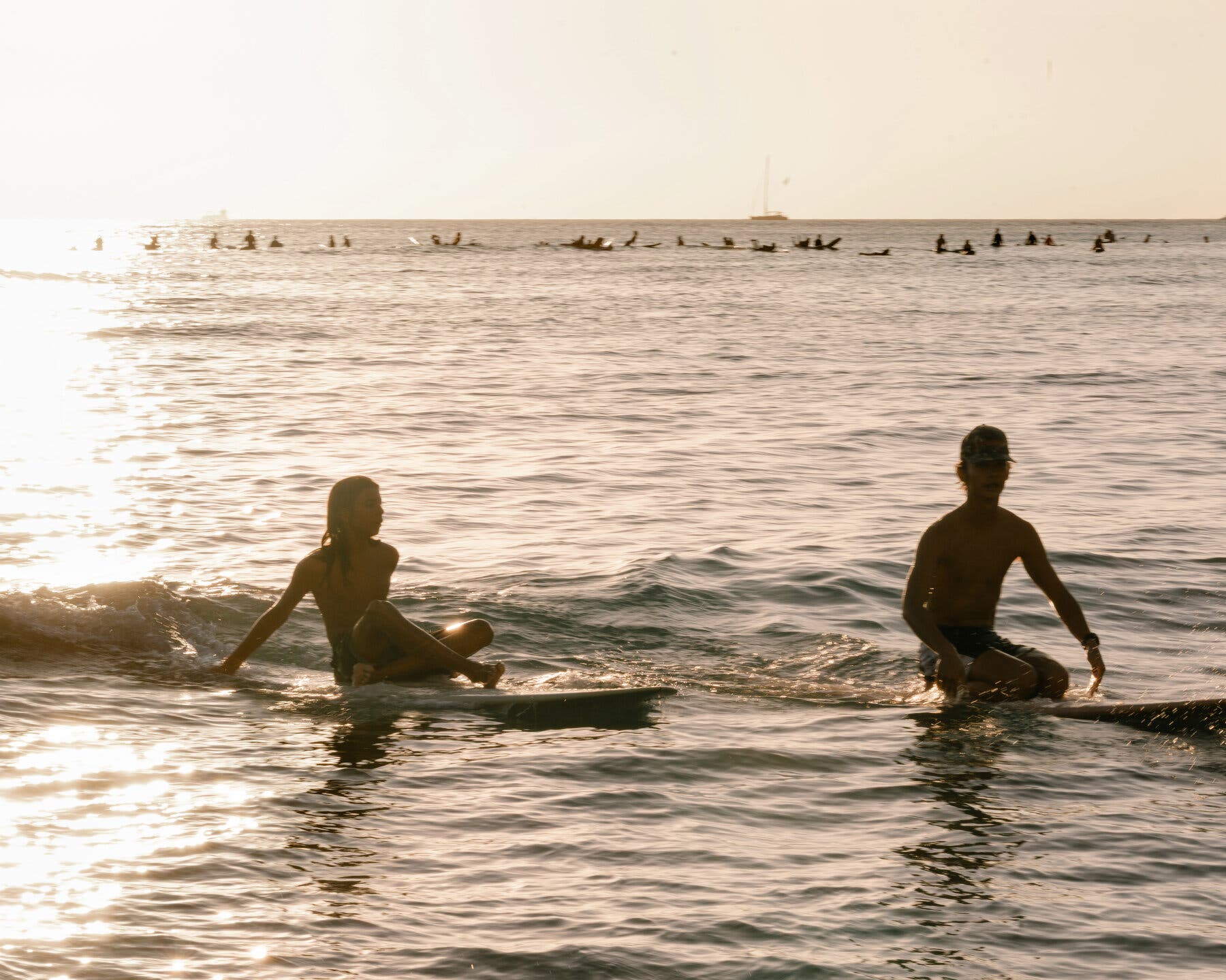 2026-02-28 ¡27trav-cheapskate-honolulu-pgvh-mobileMasterAt3x.jpgTwo people sit on surfboards in the water. Many others are on surfboards in the distant background.