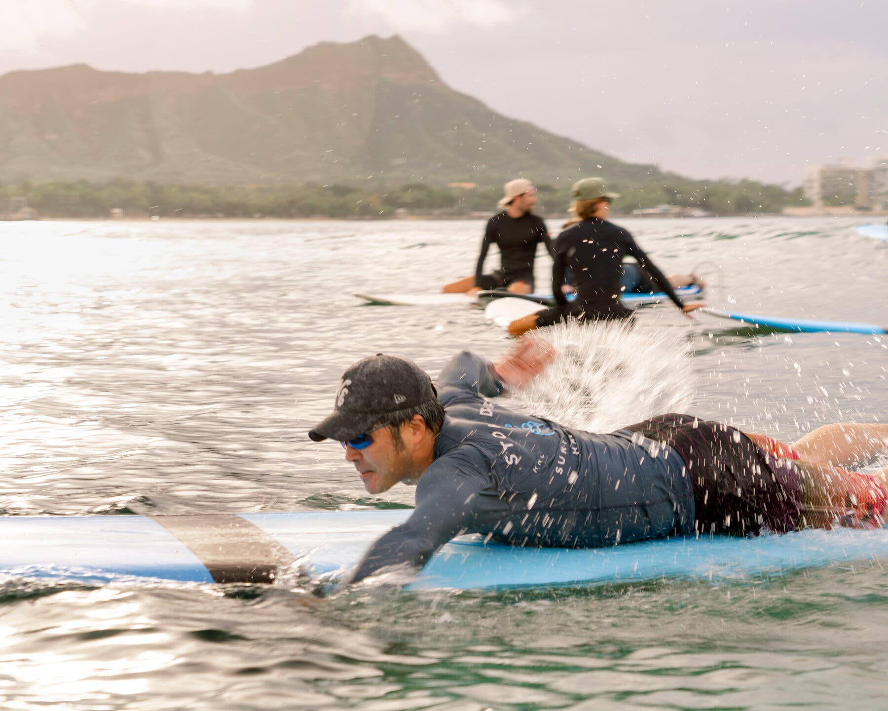 2026-02-28 ¡27trav-cheapskate-honolulu-zfwj-mobileMasterAt3x.jpgA person on a light blue surfboard paddles through water, creating splashes. Two other people are on surfboards in the distance, with a mountain and buildings behind them.