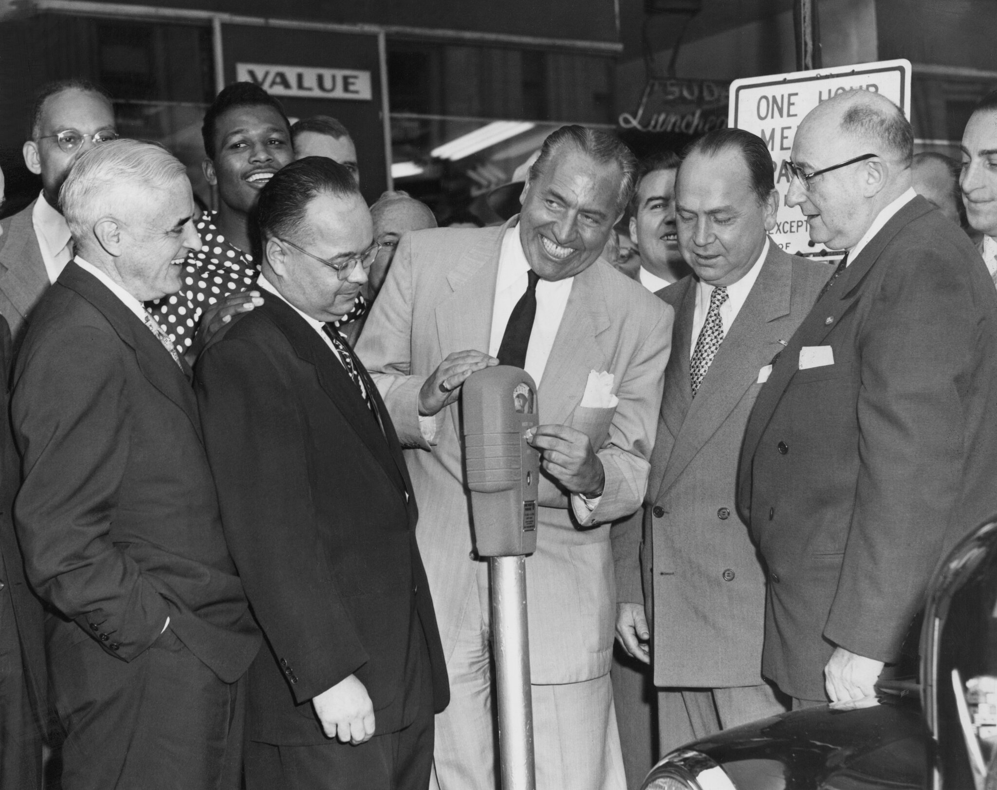 2026-06-10 ¡2026-06-10 ¡parking-timeline-04-fplg-superJumbo-v3.jpgA black-and-white 1950s photo of a man in business attire with a high-wattage grin putting a coin in a parking meter as others look on.
