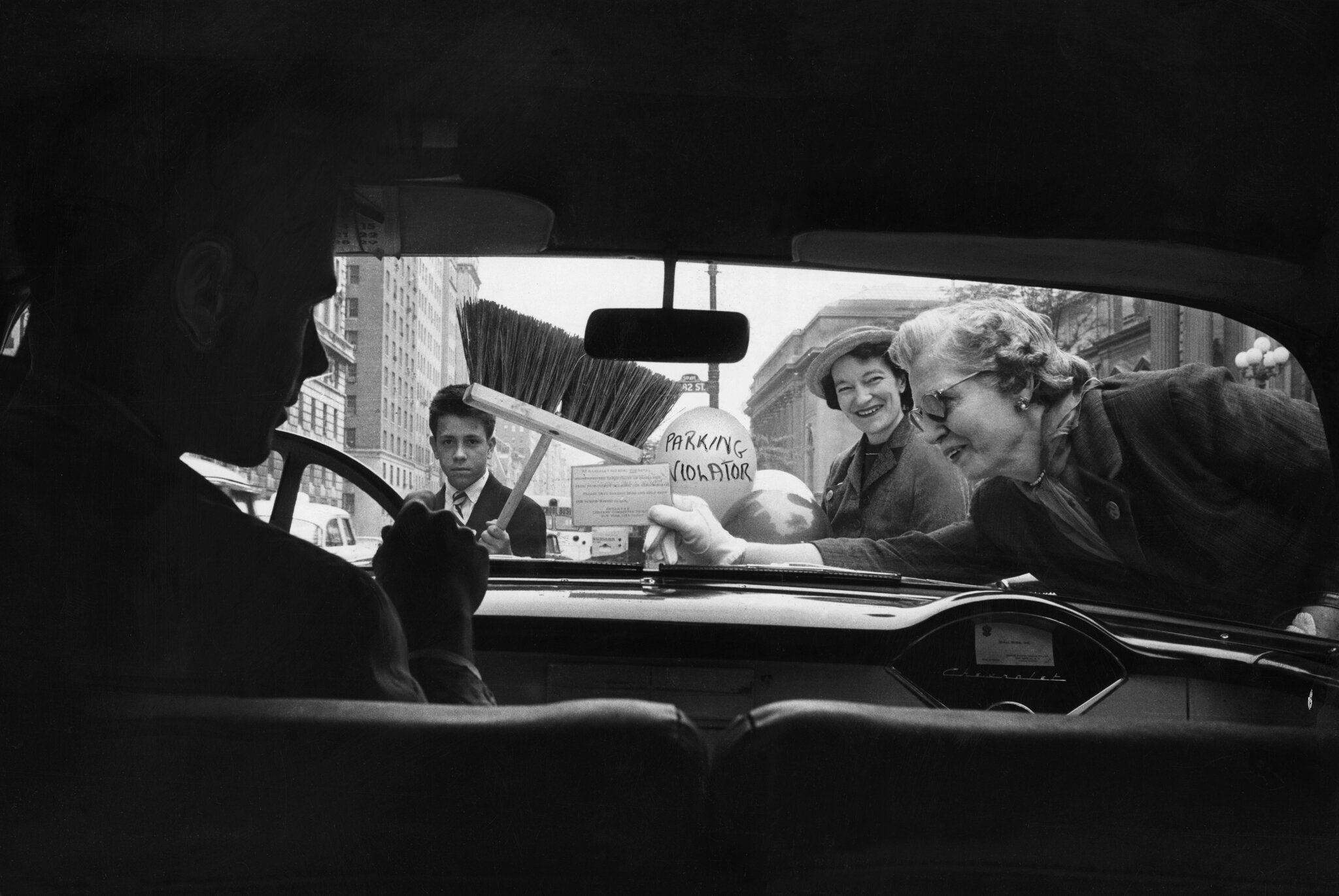2026-06-10 ¡parking-timeline-14-fplg-superJumbo.jpgA man in front of a steering wheel inside a car. Outside the car, a woman leans across the windshield holding a sign. Another woman holds a balloon that says “parking violater” and a boy stands with a broom.