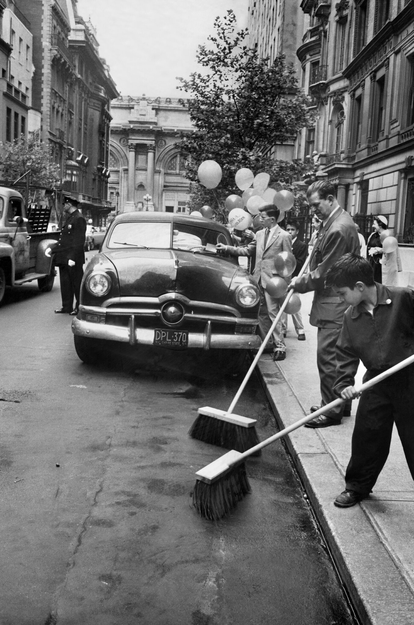 2026-06-10 ¡parking-timeline-23-fplg-superJumbo-v3.jpgA black-and-white photo from the 1950s of people with brooms performatively sweeping a street in front of a parked car, with balloons in the background.