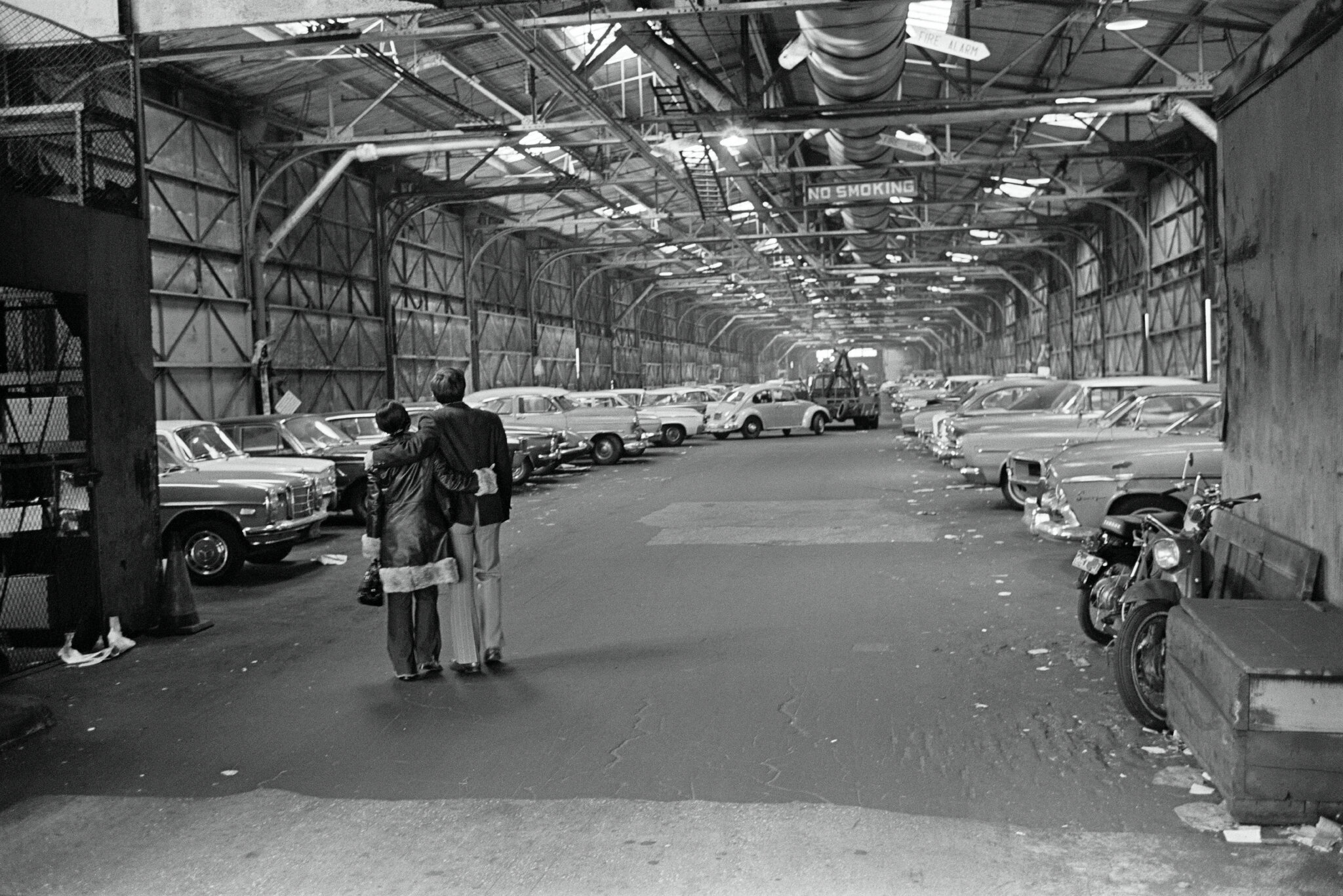 2026-06-10 ¡parking-timeline-vbcf-superJumbo.jpgA black-and-white photo from 1969 of a man and woman with their backs to the camera looking at a large covered garage with dozens of cars in it.